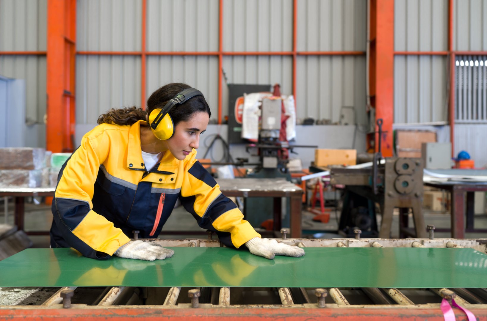 Young female hispanic labor dressed in polyester jacket uniform and ear muff working in metal sheet manufacture. Various kind of industrial machine are in the working area.