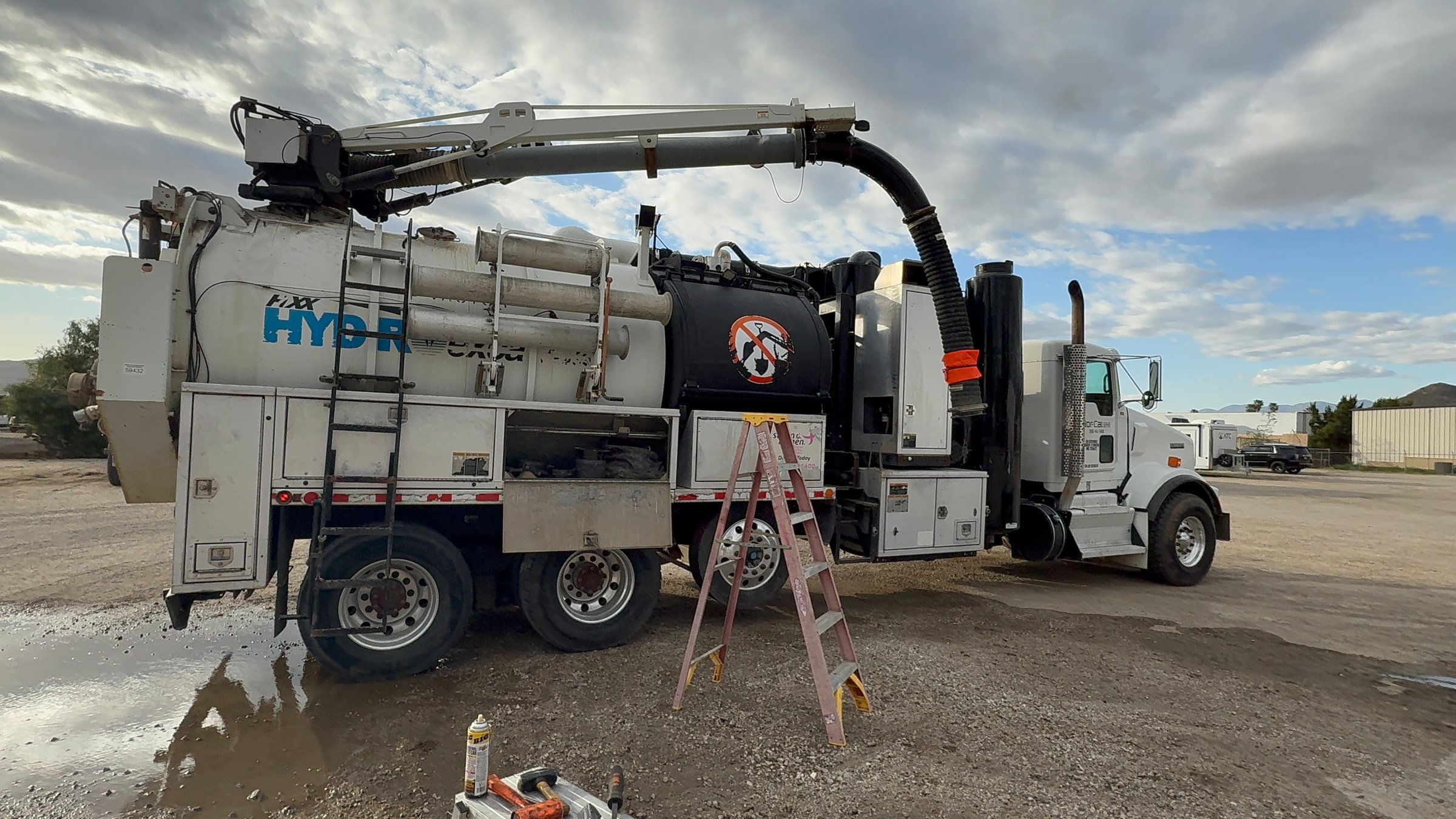 A large vacuum truck with HYDR printed on its side, parked with a ladder leaning against it, under a cloudy sky.