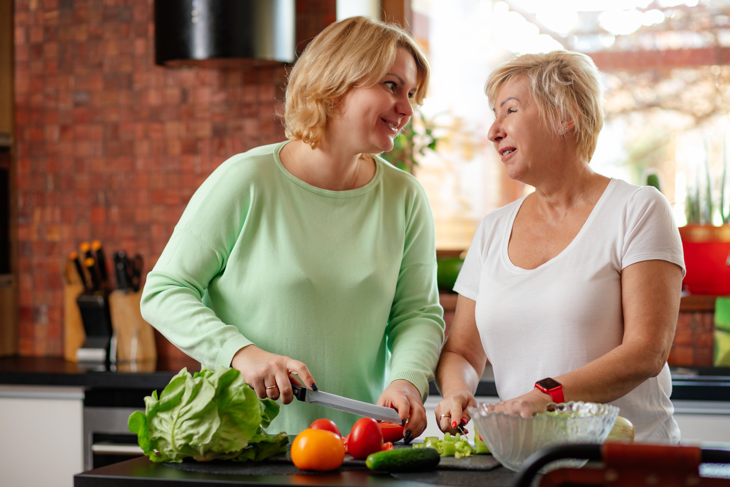 Blonde smiling senior woman and daughter help cook dinner together in light kitchen. Family grandmother teach housekeeping prepare healthy food vegetable lunch two person lifestyle.
