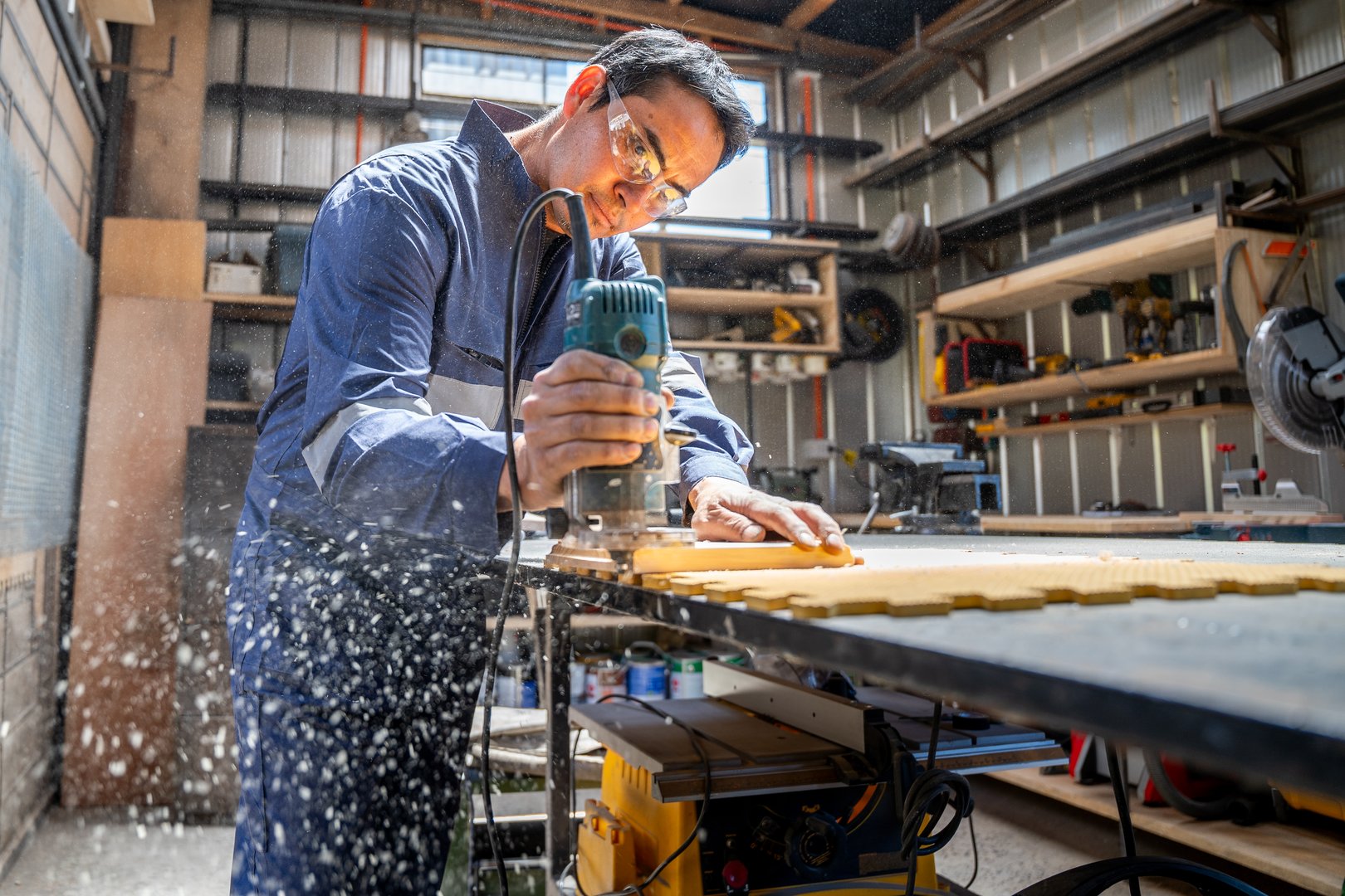 A focused man uses a router on wood, creating precise cuts while surrounded by tools in a well-organized workshop.