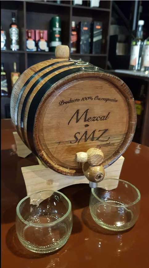 Wooden mezcal barrel on a stand with two glass cups in front, displayed in a bar setting with bottles in the background.