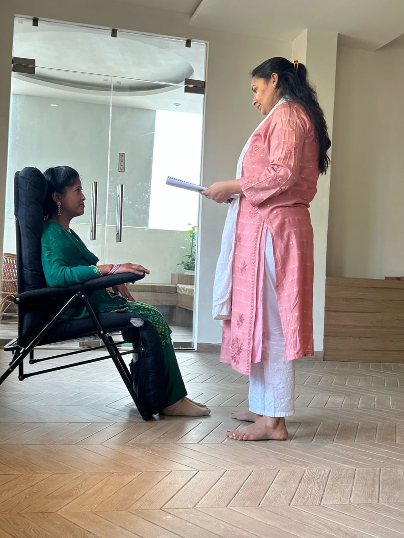 Two women having a conversation indoors; one seated in a chair, the other standing with a clipboard.