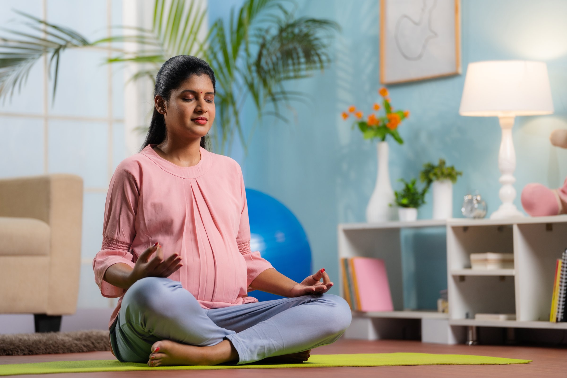 Indian peaceful pregnant woman in lotus pose doing meditation on yoga mat home - concept of mindfulness, relaxation and Self-care during pregnancy.