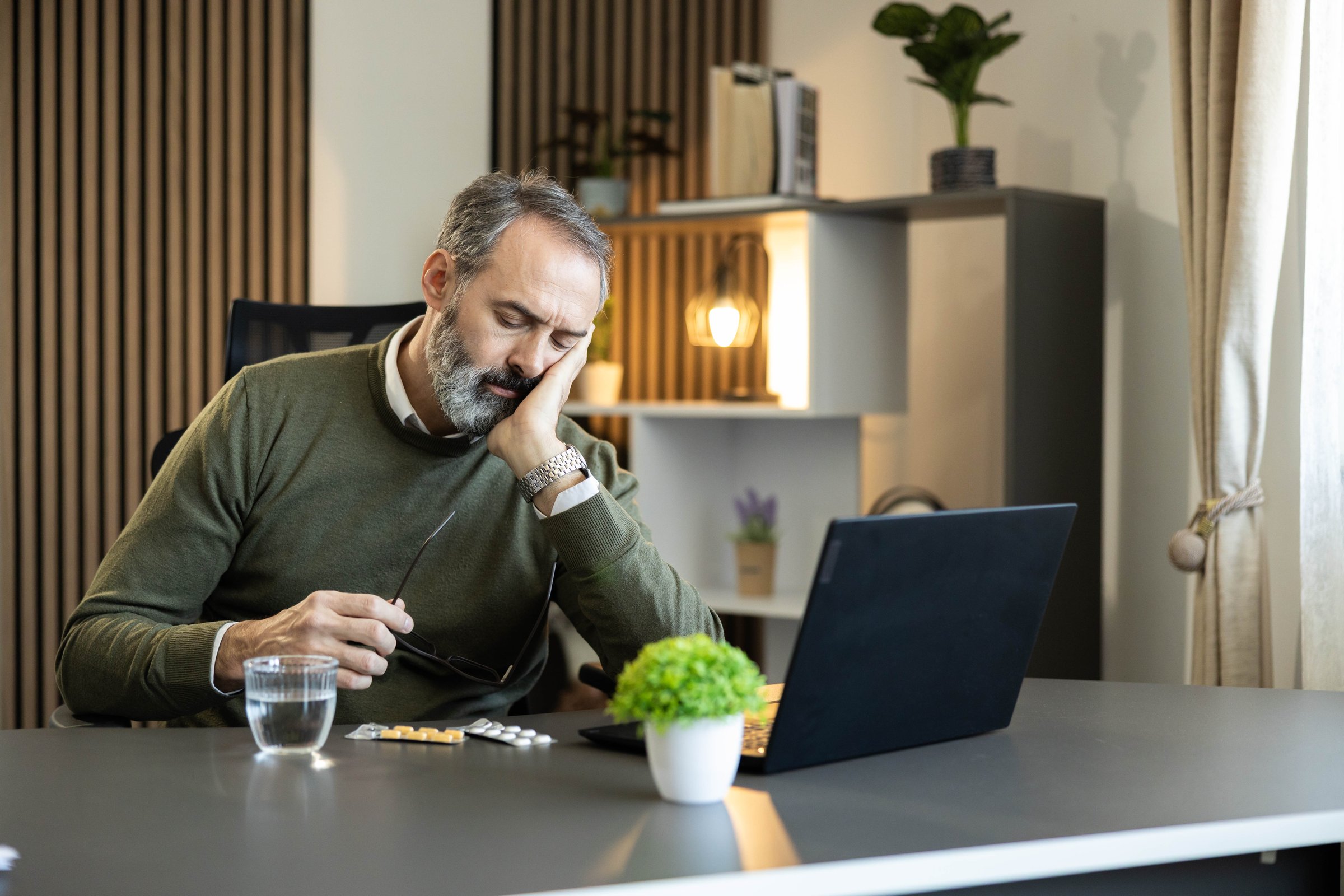 Stressed mature man experiencing exhaustion and overwork symptoms, holding glasses in hands with medicine and water on desk