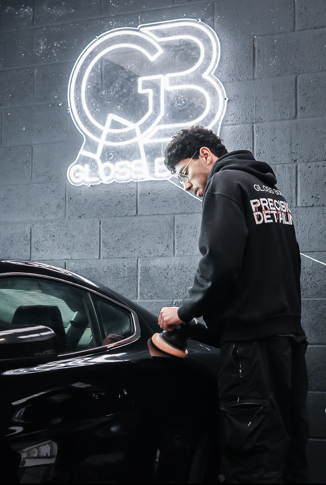 Person polishing a black car in a garage with a neon GLOSSBE sign on the wall.