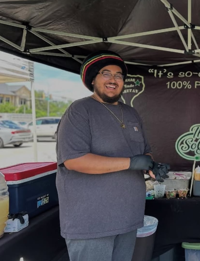 Person smiling at an outdoor food stall, wearing a black T-shirt and hat, with beverages and condiments on the table.