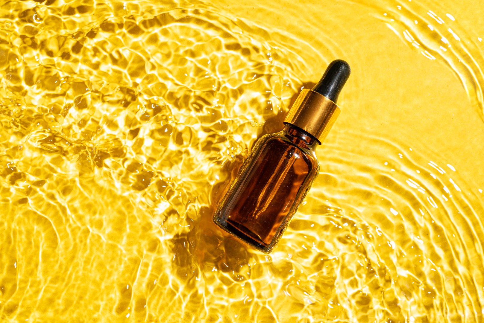 Dropper bottle with retinol serum in bright yellow water background. Creative template for design. Vitamin C serum on waved water surface.