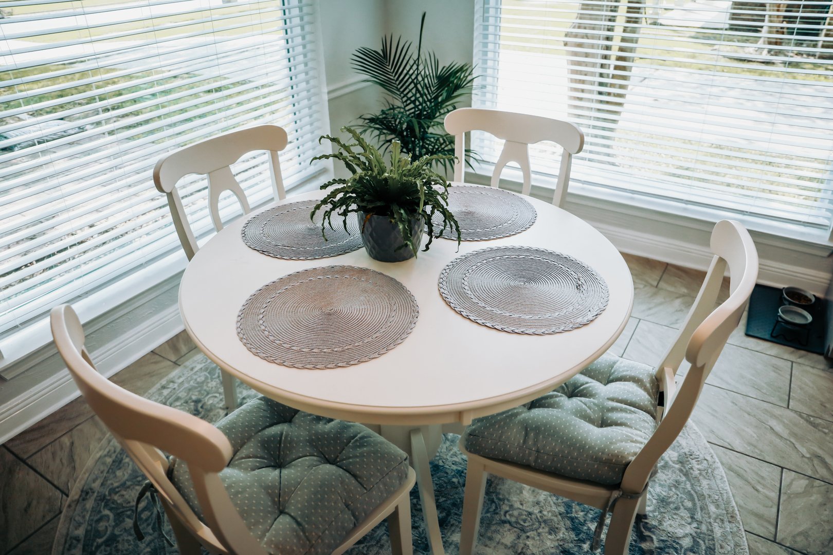 Close up of a small table and four chairs in an eat in kitchen with a tile floor, a rug, plant, and placemats.