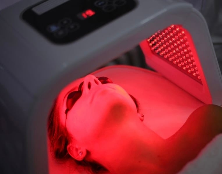 Person receiving red light therapy, wearing protective goggles, under a device emitting red light on the face.