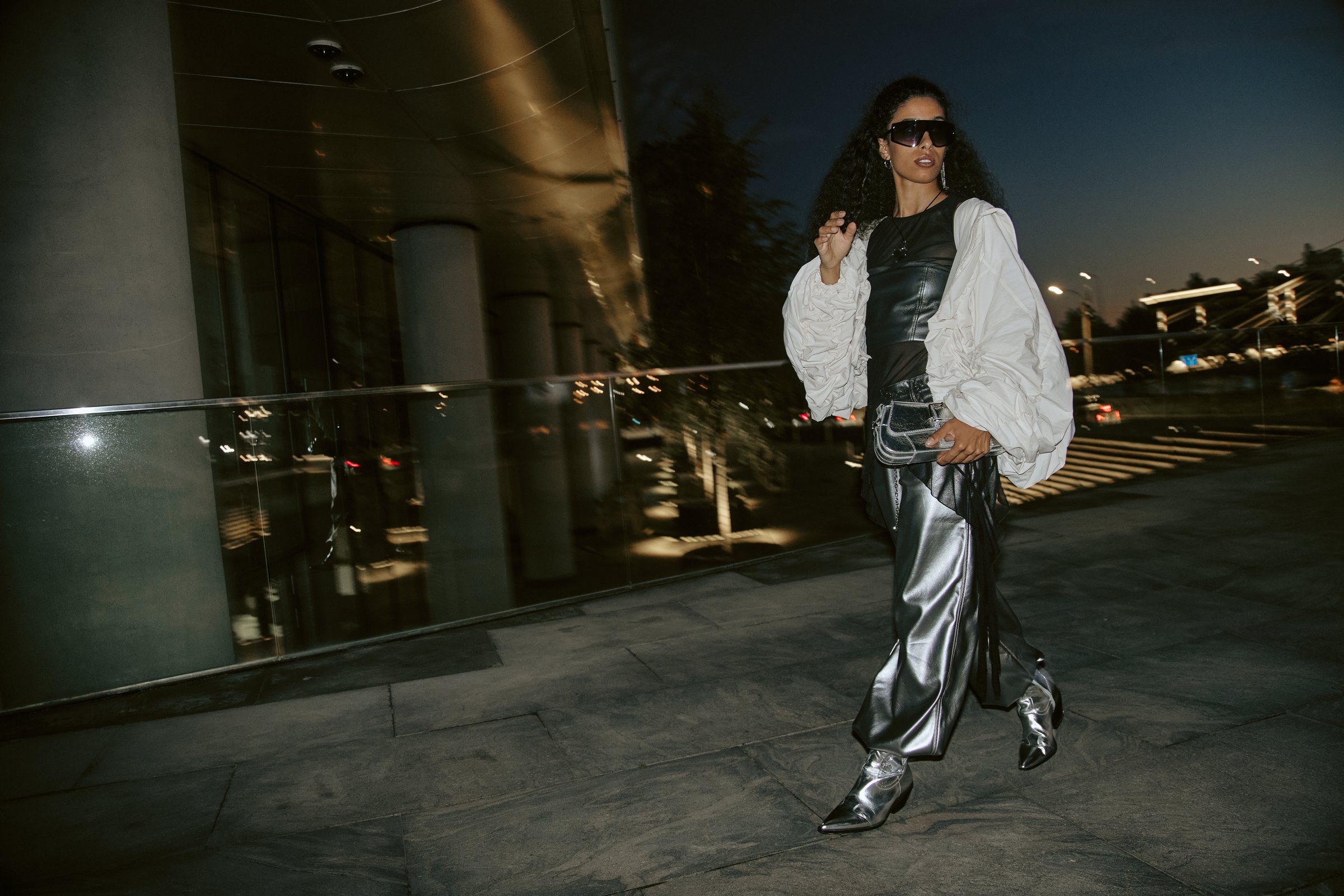 Full shot of Middle Eastern female celebrity wearing silver pants paired with cowboy boots and purse, while walking through camera lights in night city showcasing cool metallic outfit, copy space