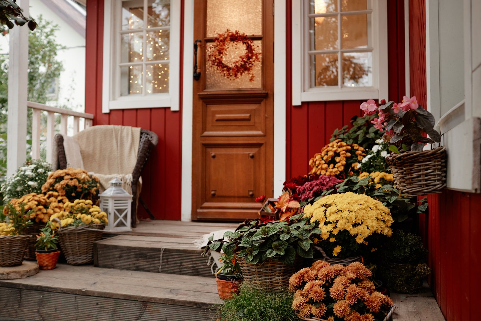 Front porch of village house decorated with autumn flowers and plants, wooden steps surrounded by baskets of blooming chrysanthemums, cozy rural setting with seasonal wreath on door