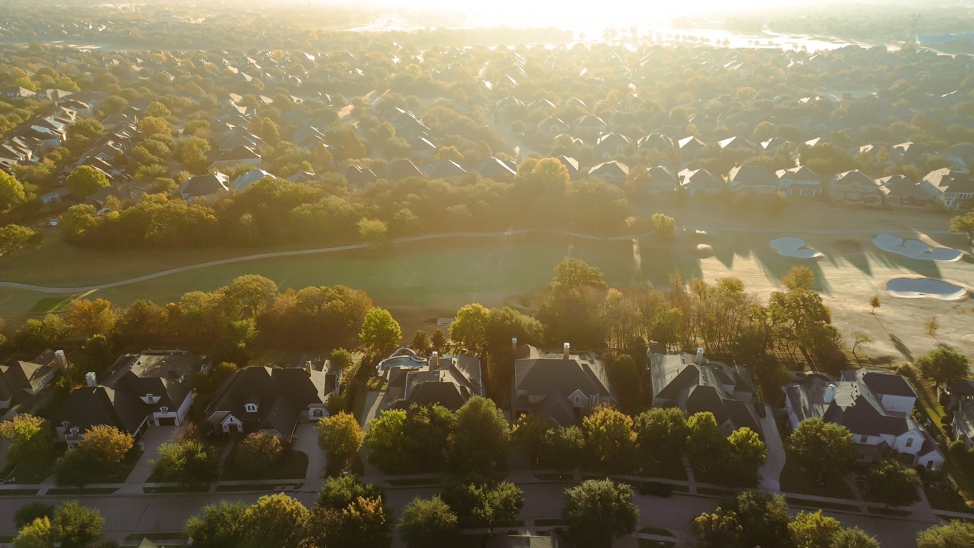 Sunset over community golf course and upscale residential homes in Lantana, Texas, sand bunkers, walking paths, green space, captured in soft light with tree shadows and hazy suburban backdrop. USA