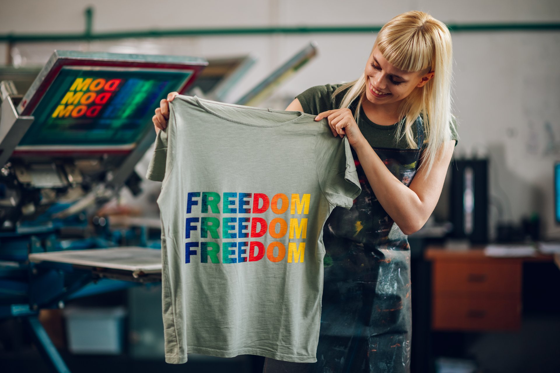 Smiling female printing workshop worker standing at facility with silkscreen printed t-shirt and showing it at the camera and promoting lgbtq equality. Print shop worker with finished product in hands