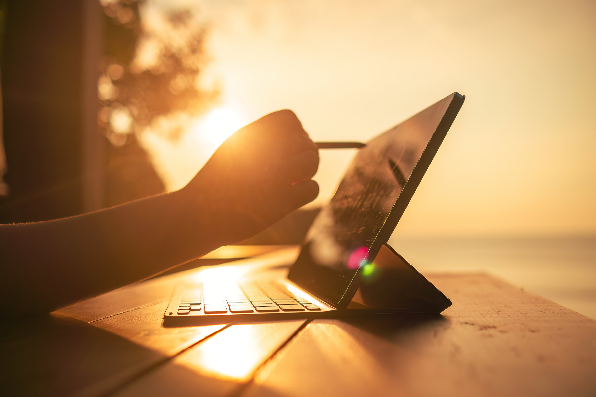 Close-up of a person's hand using a stylus on a tablet computer, at a wooden table, with a sunset in the background. The sun is golden, creating a warm and peaceful atmosphere. Remote work concept.