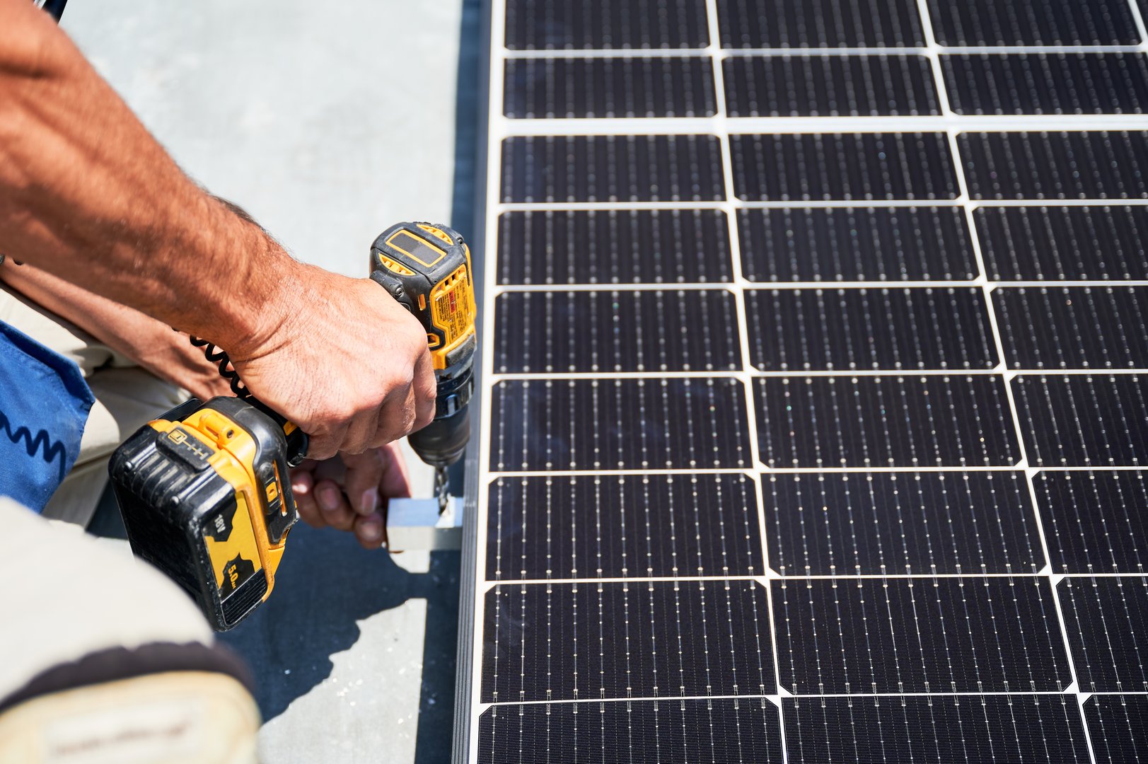 Worker building photovoltaic solar panel system on metal rooftop of house. Close up man technician installing solar module with help of electric drill outdoors. Alternative energy generation concept.