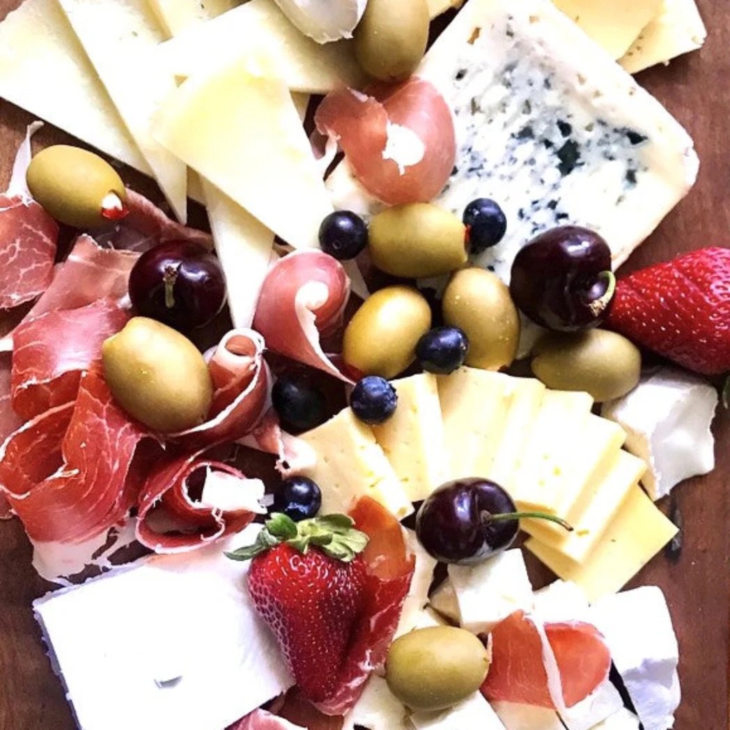 Charcuterie board with cheeses, prosciutto, olives, cherries, strawberries, and blueberries on a wooden surface.