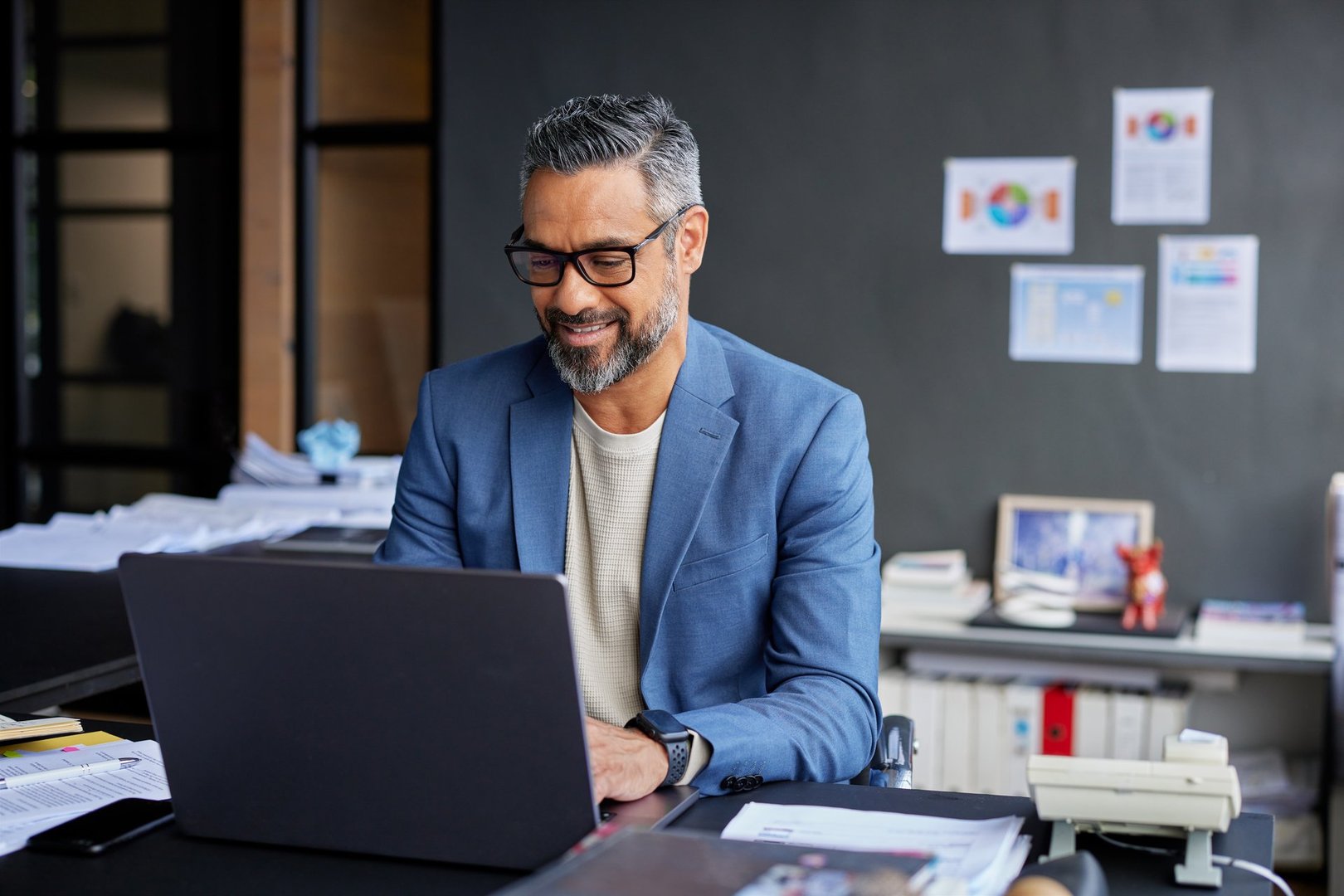 Mid adult businssman smiling while working on laptop in his office. Confident professional mixed race business man working in modern office while sitting at desk. Mature businessman engaged in online work while wearing eyeglasses.
