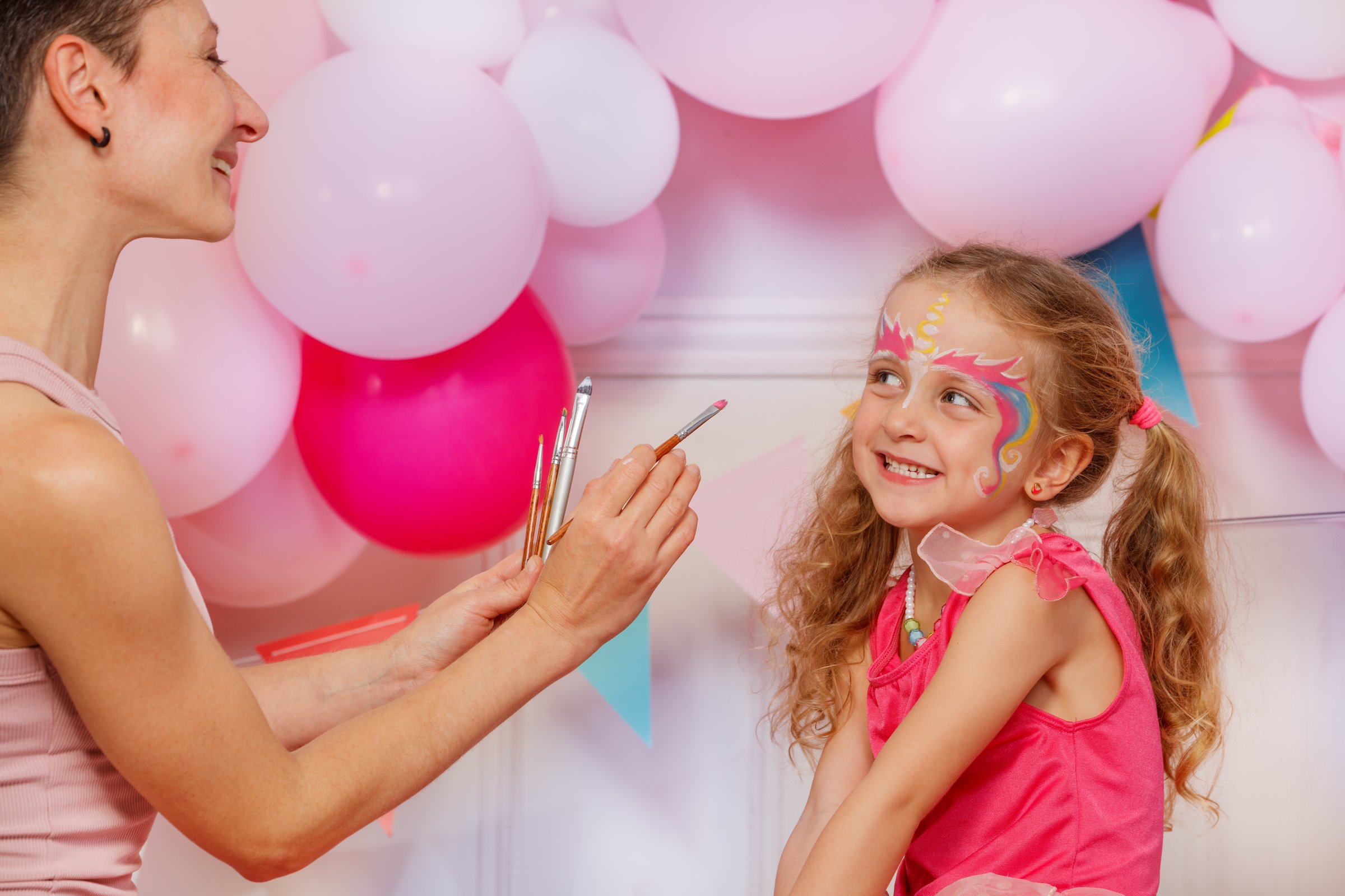 In front of pink balloons, a woman is doing fantasy makeup on a child's smiling face with bright hue colors