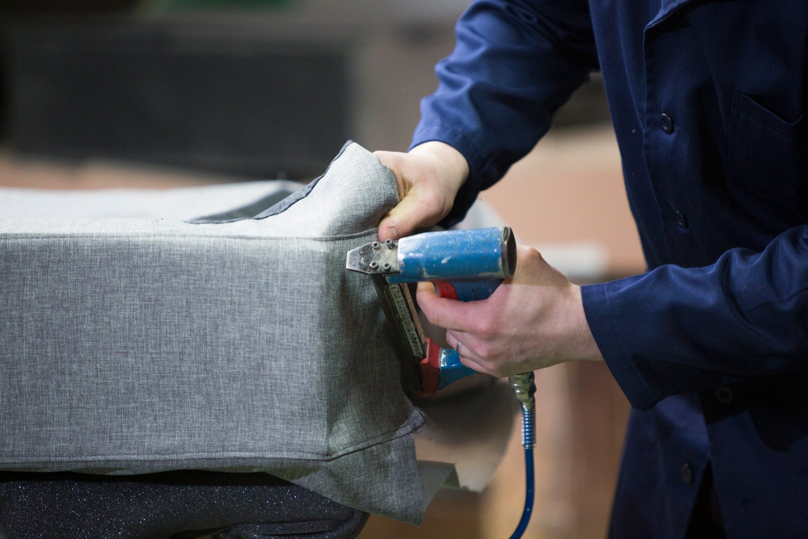 Process of furniture production. A young man in a dark coat who puts together one part of the sofa with a stapler on a working desk. He has a black safety gloves.