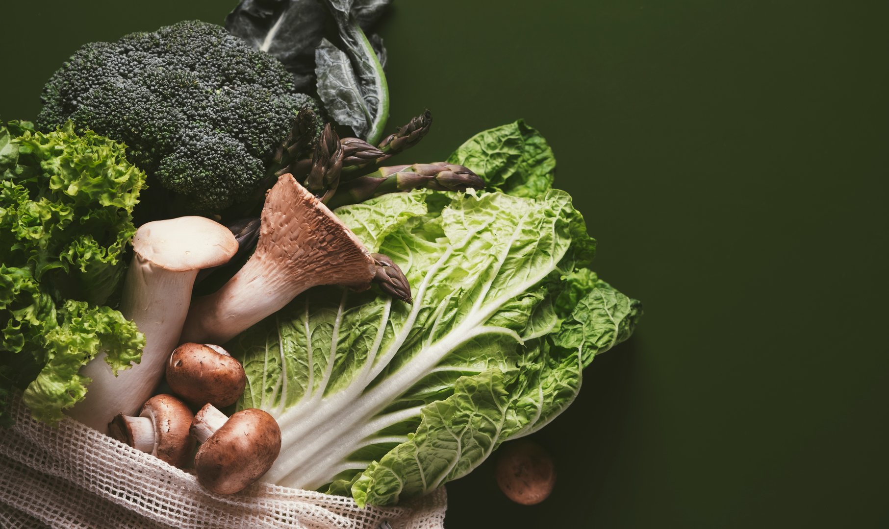 Vibrant green vegetables and herbs fill a reusable bag against a green backdrop, perfect for creating nutritious meals from a local market.