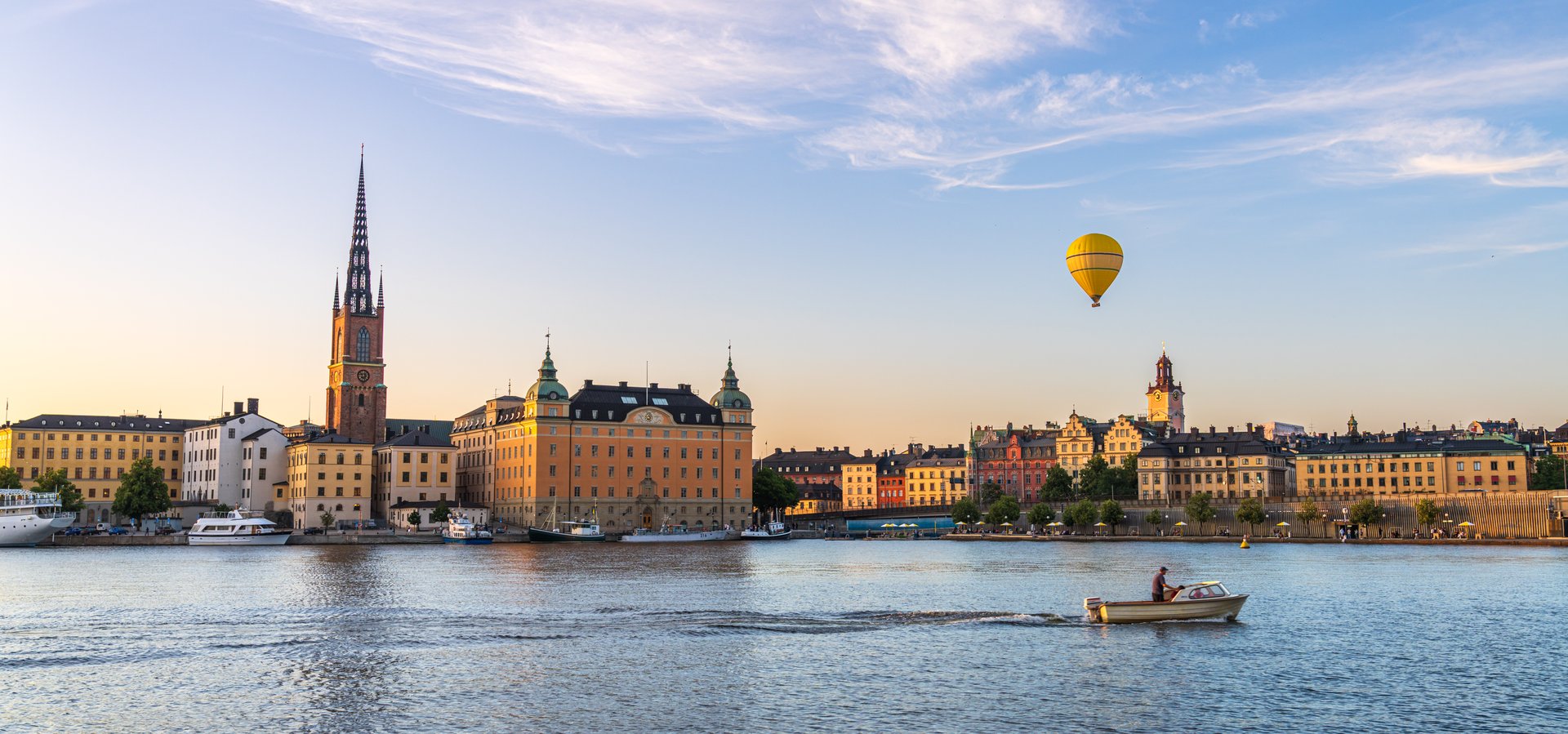 View of Riddarholmen's silhouette against the water, a yellow hot air balloon floats above the buildings, boat passes by, Stockholm, Sweden.