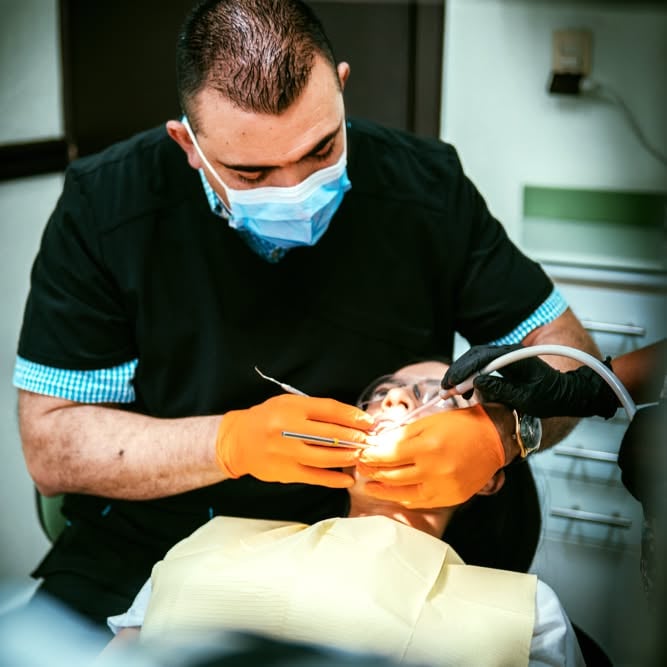 Dentist wearing orange gloves and a mask performs a dental procedure on a patient reclined in a chair.