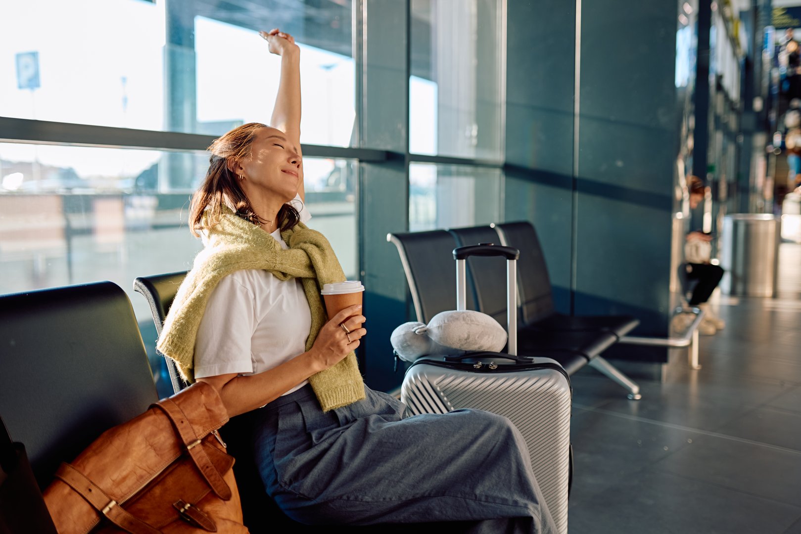 Happy woman relaxing at airport, drinking coffee, stretching arm, and waiting for her flight with luggage in sunny terminal
