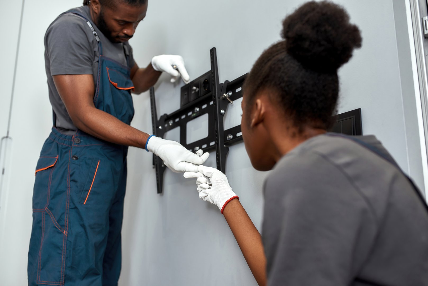 Repair and relocation concept. African American woman and man holding large flat screen TV, hanging it on grey wall in modern appartment. Light grey living room with wooden floor.