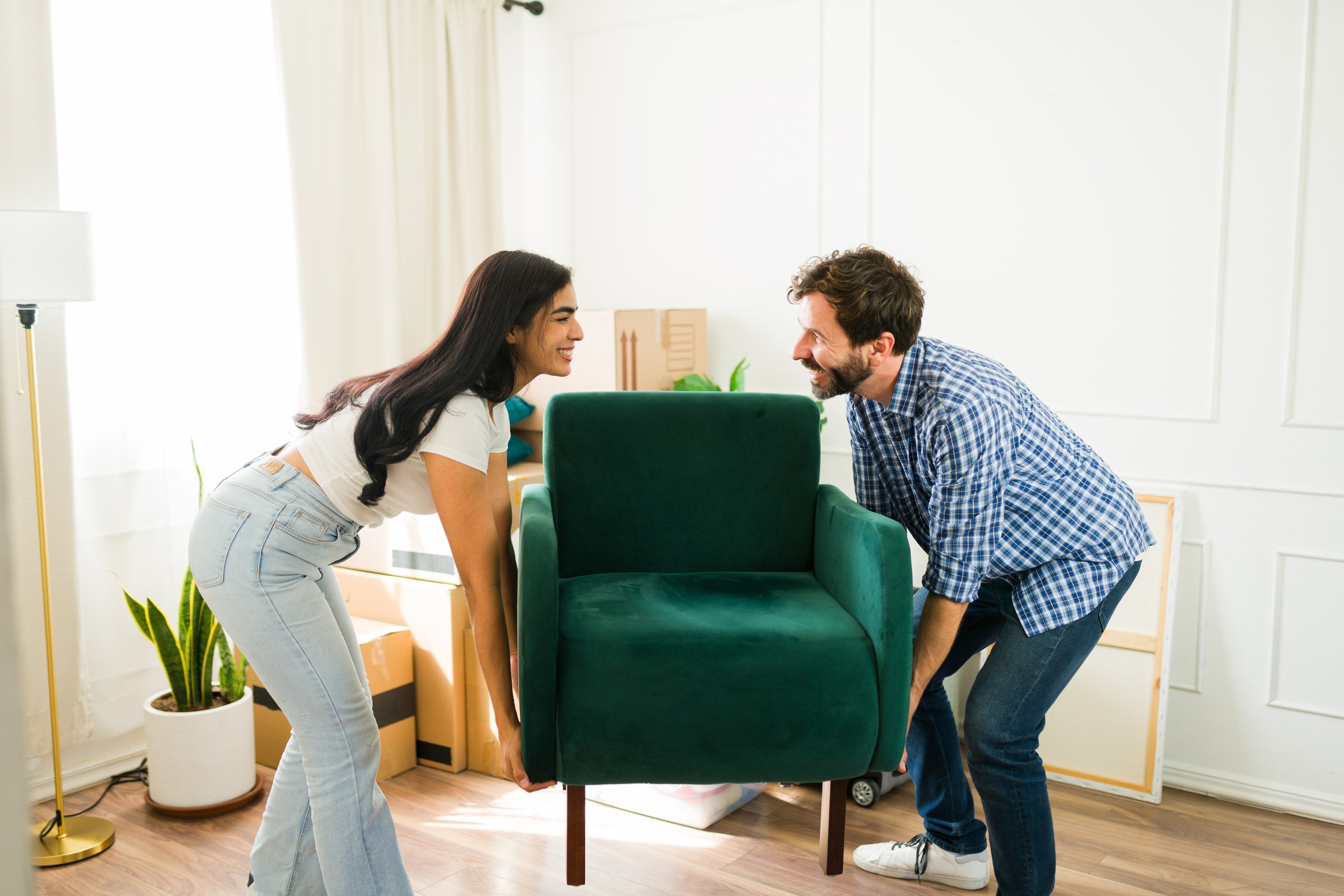 Young couple is carrying an armchair together while smiling and arranging furniture in their new home