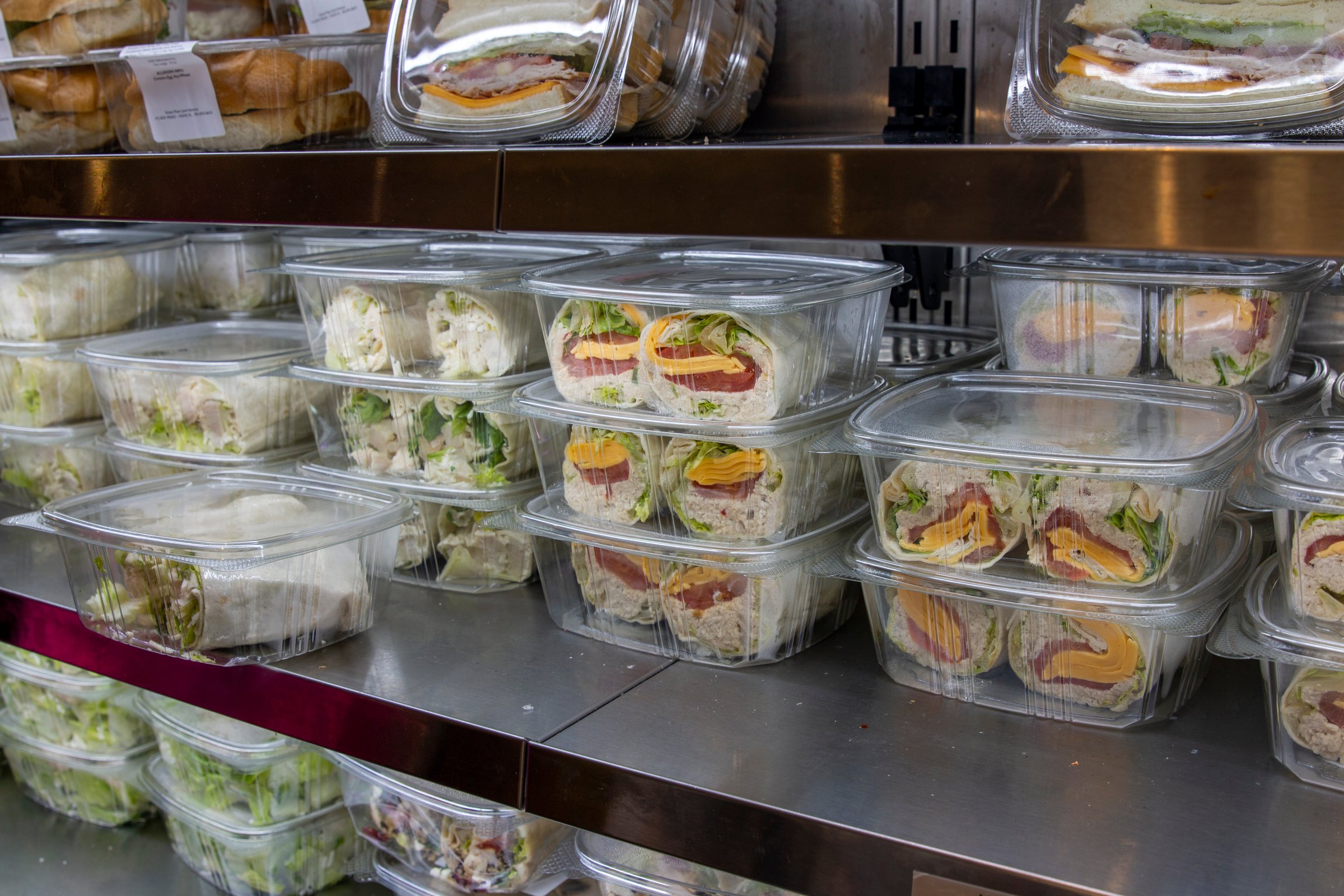 A variety of neatly arranged sandwich and salad containers are prominently displayed on a stainless steel shelf, ready for lunchtime customers in an urban deli environment.