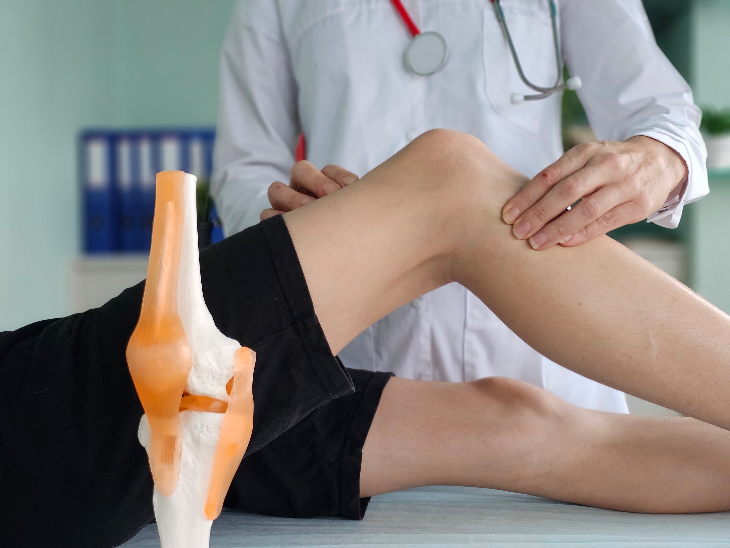 A physical therapist examines the knee of a teenager sitting on an examination table in a clinic office