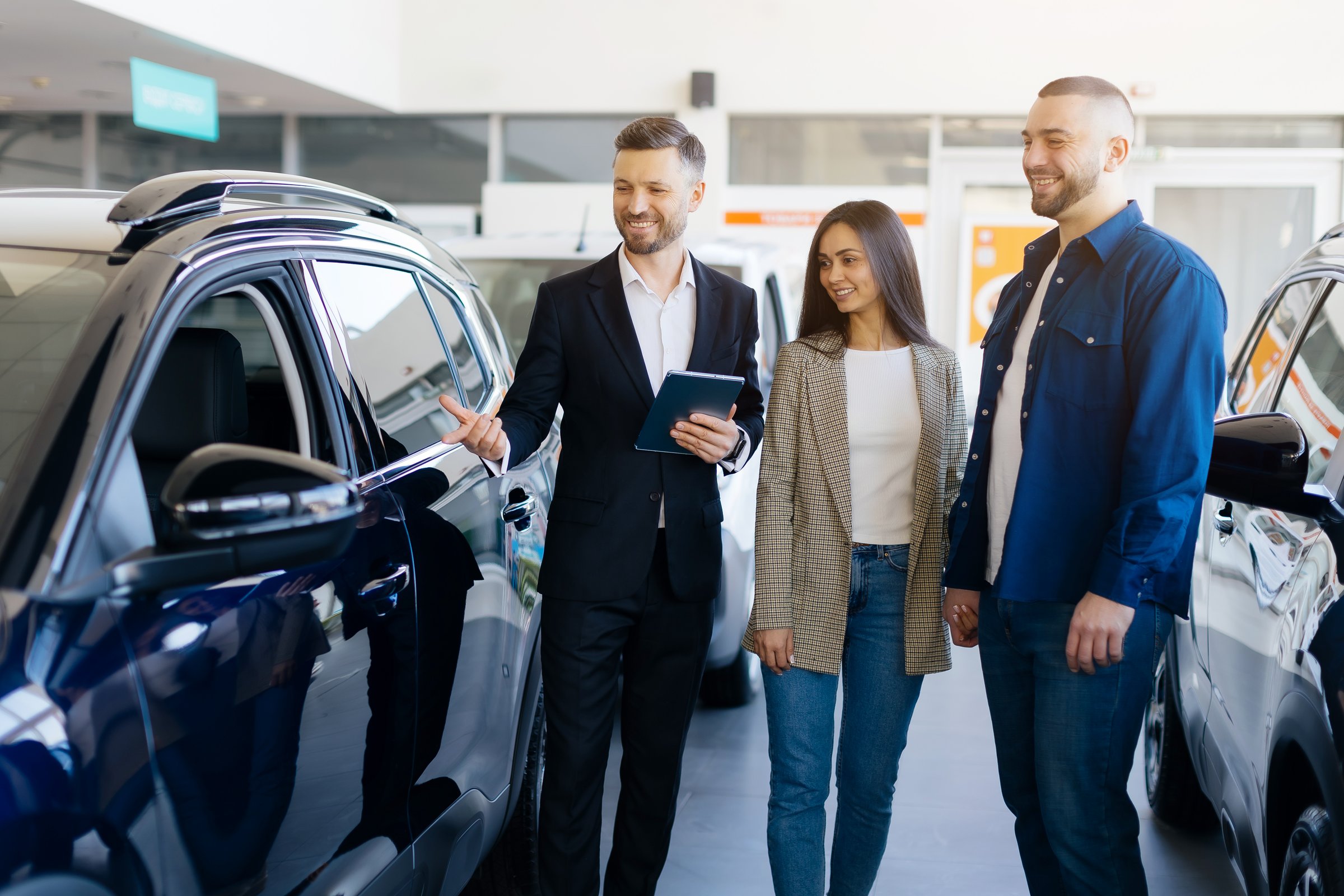 Family Buying Car. Young Spouses Purchasing New Vehicle In Dealership Center, Happy Millennial Couple Discussing Model Of Auto With Smiling Salesman In Modern Showroom