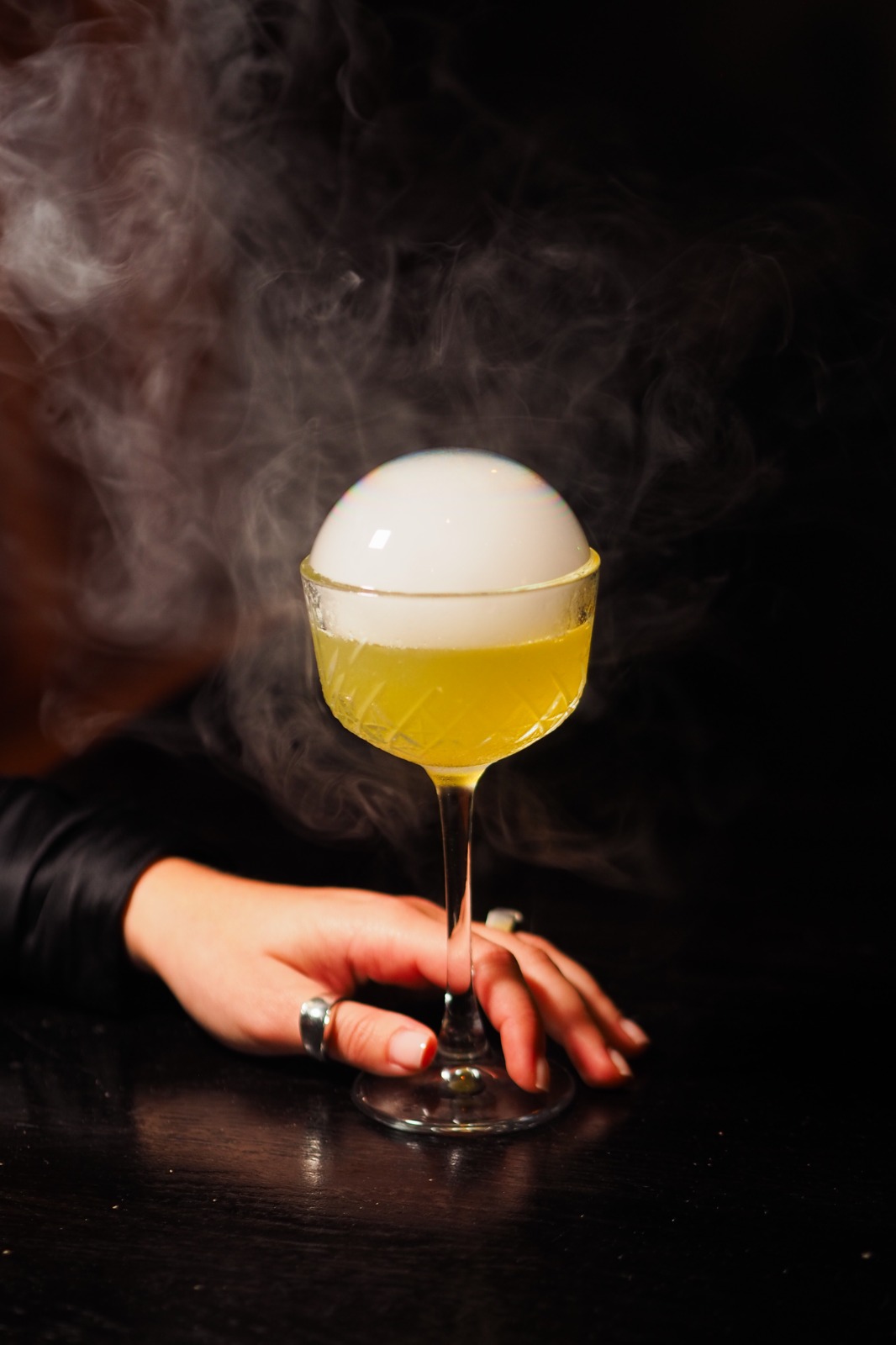 A hand holding a cocktail with a smoke-filled bubble on top, in a glass on a dark background.