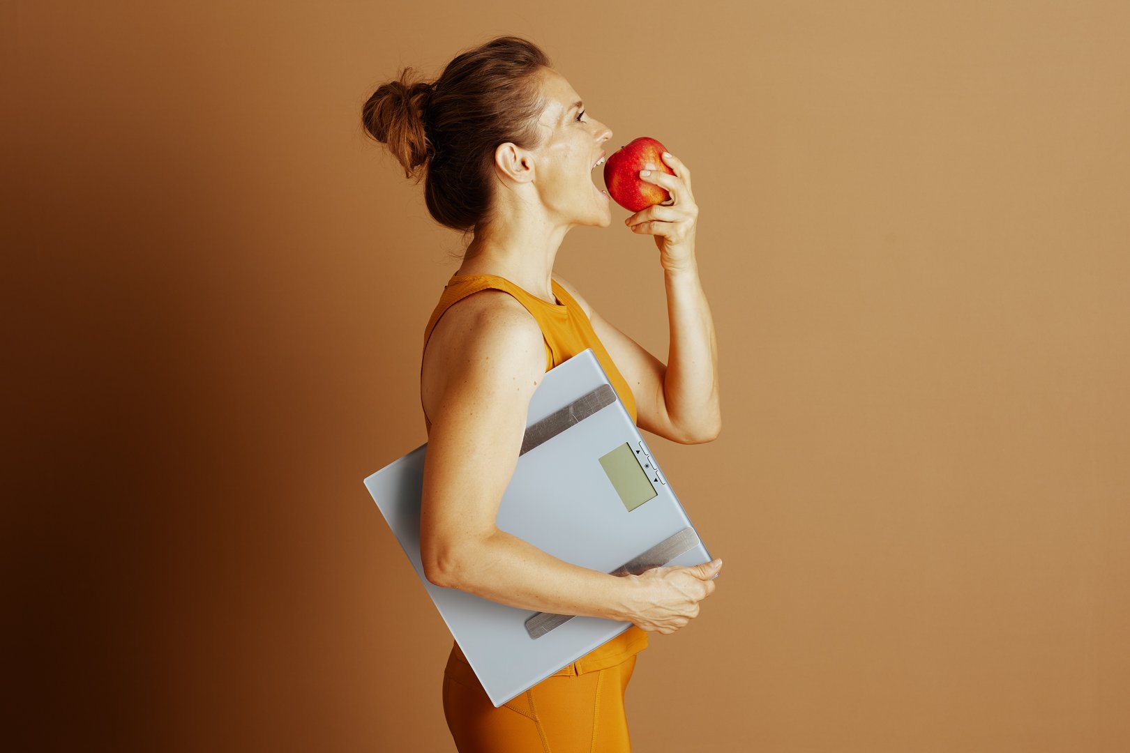 A mid-adult woman, in profile, holds a red apple to her mouth and a digital weight scale, symbolizing a balanced approach to healthy eating, diet, and mindful weight management for overall wellness.