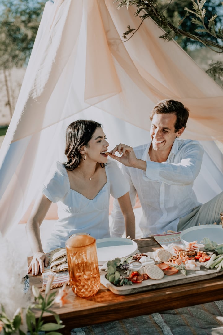 Couple enjoying a picnic in front of a tent, smiling as the man offers food to the woman. The table is set with snacks and drinks.