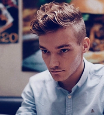 Young man with styled hair, wearing a light blue shirt, looking serious in a softly lit indoor setting.