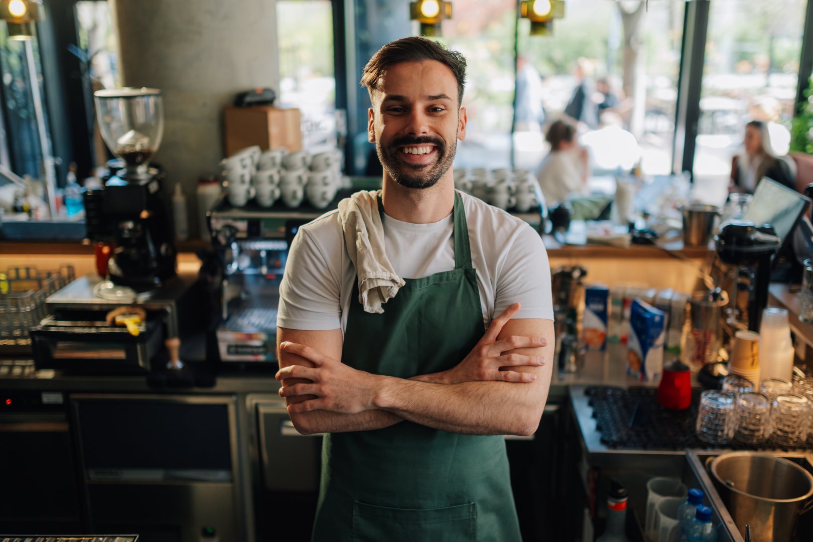 Young barista smiling confidently with crossed arms, proudly representing his coffee shop and showcasing his passion for the craft