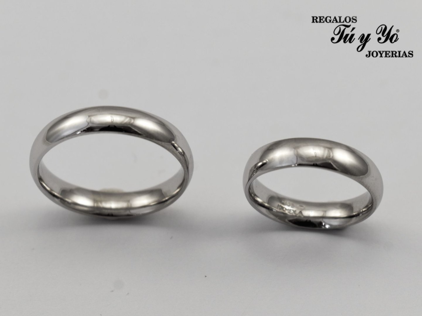 Two silver wedding bands with reflective surfaces, displayed side by side against a neutral background; brand text in corner.