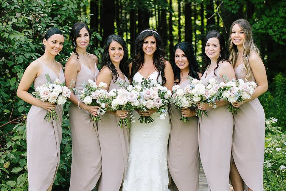Bride in white gown poses with six bridesmaids in taupe dresses, holding flower bouquets, in a wooded outdoor setting.