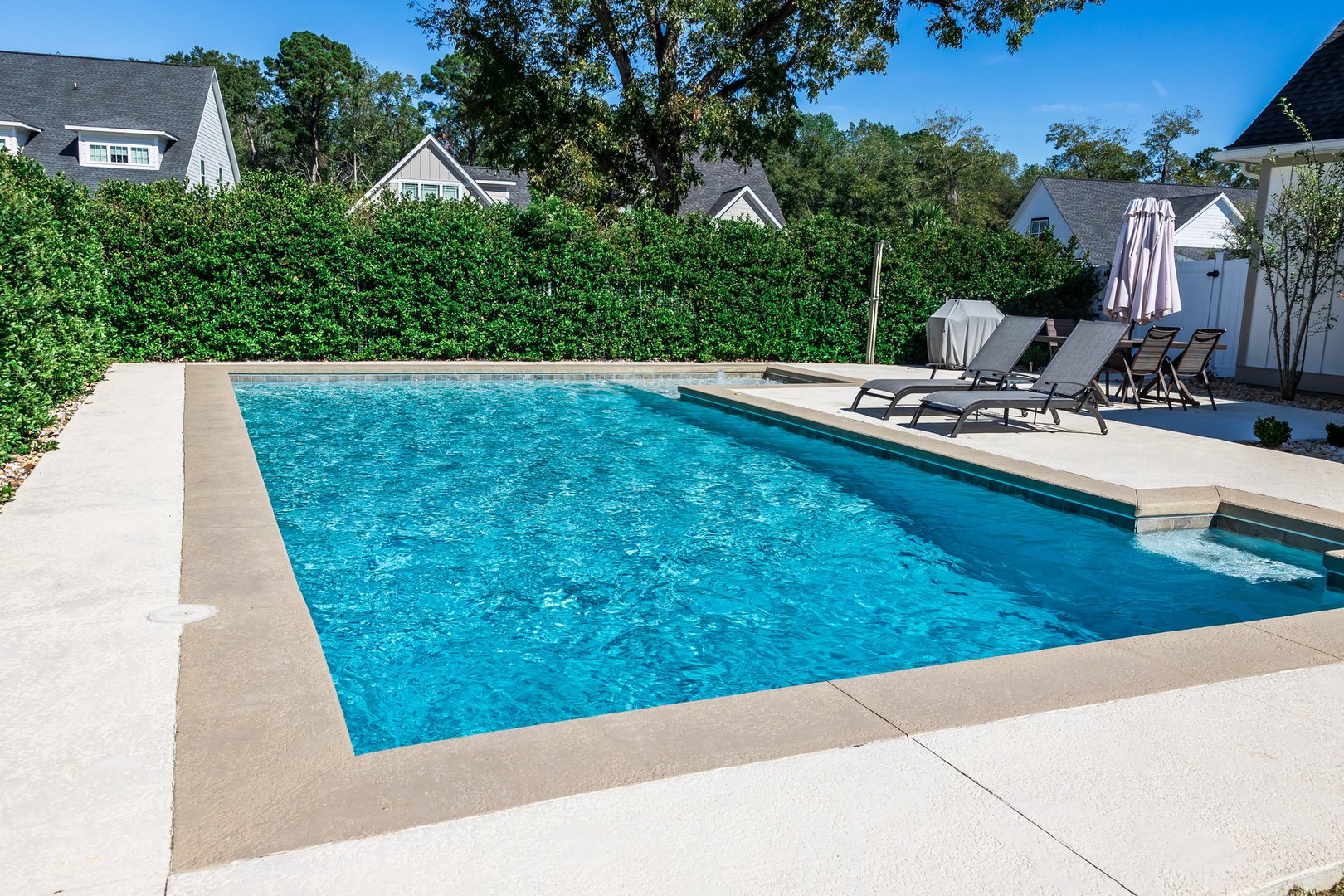 A rectangular new swimming pool with tan concrete edges in the fenced backyard of a new construction house with privacy hedges.