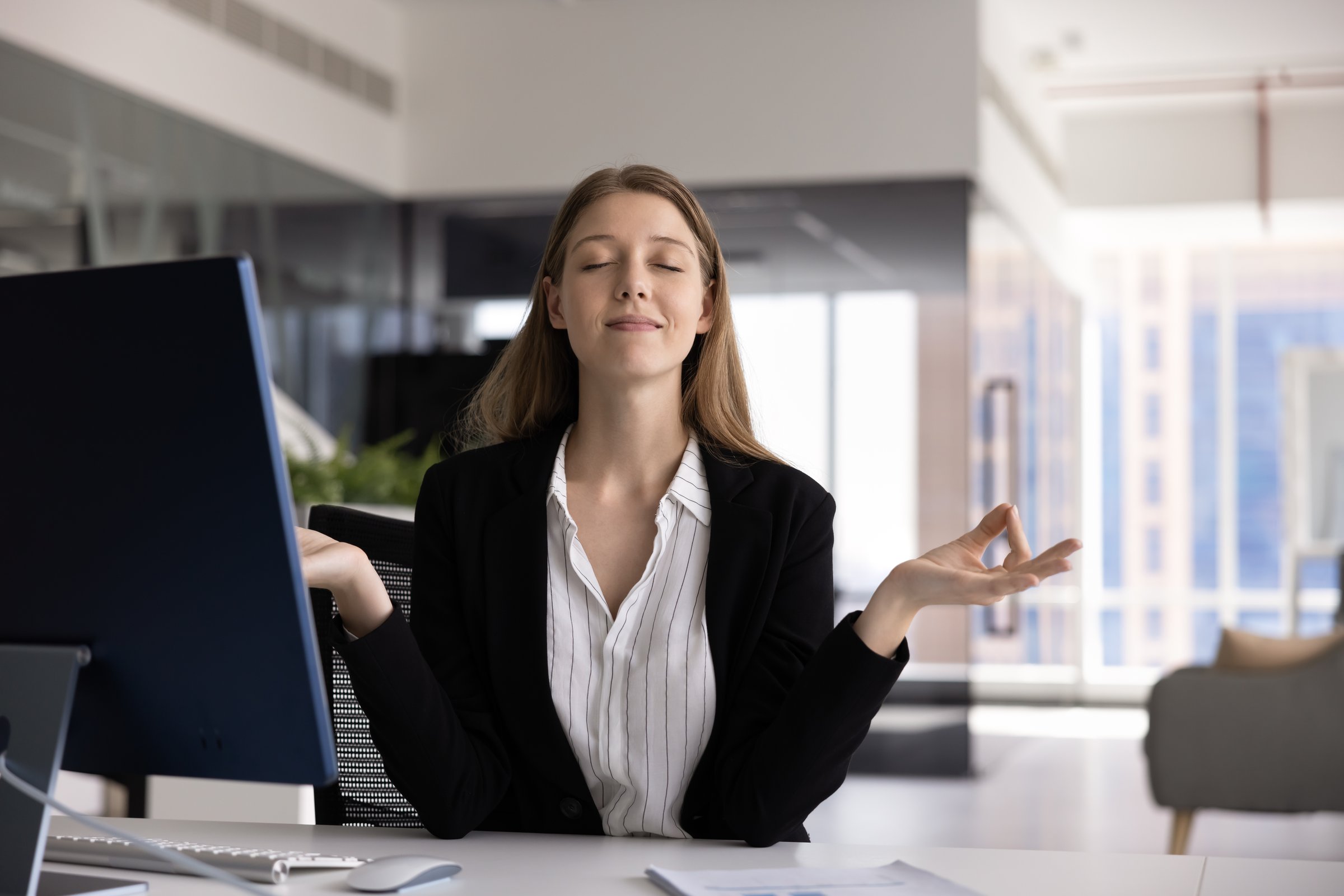 Positive calm 20s manager girl meditating at workplace, sitting at computer with zen hands and closed eyes, smiling, practicing relaxation, mindfulness, getting emotional harmony, peace, stress relief