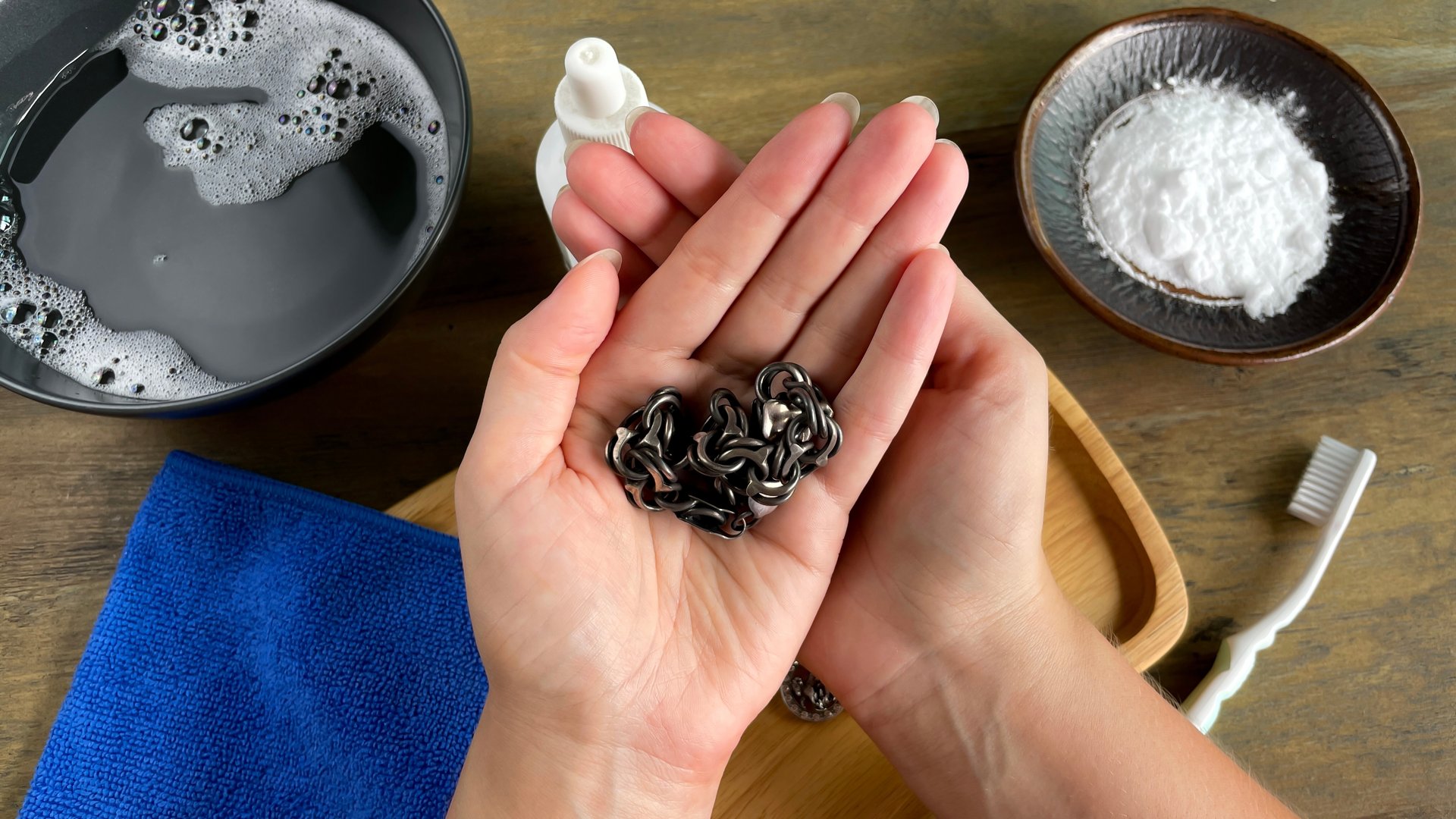 A woman's hands hold tarnished silver. On the table are soapy water and jewelry care products.