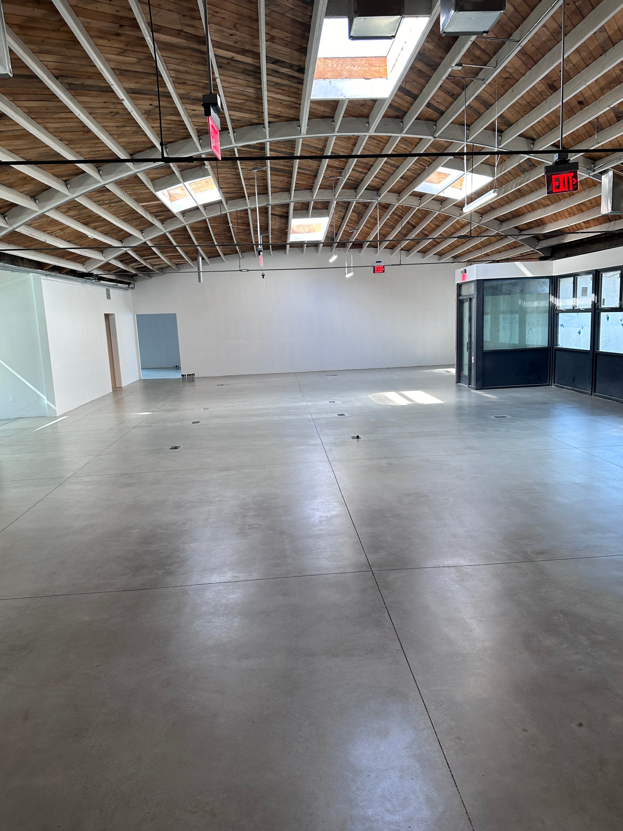 Empty industrial space with concrete floors, wooden ceiling, skylights, and glass-walled meeting room on one side.