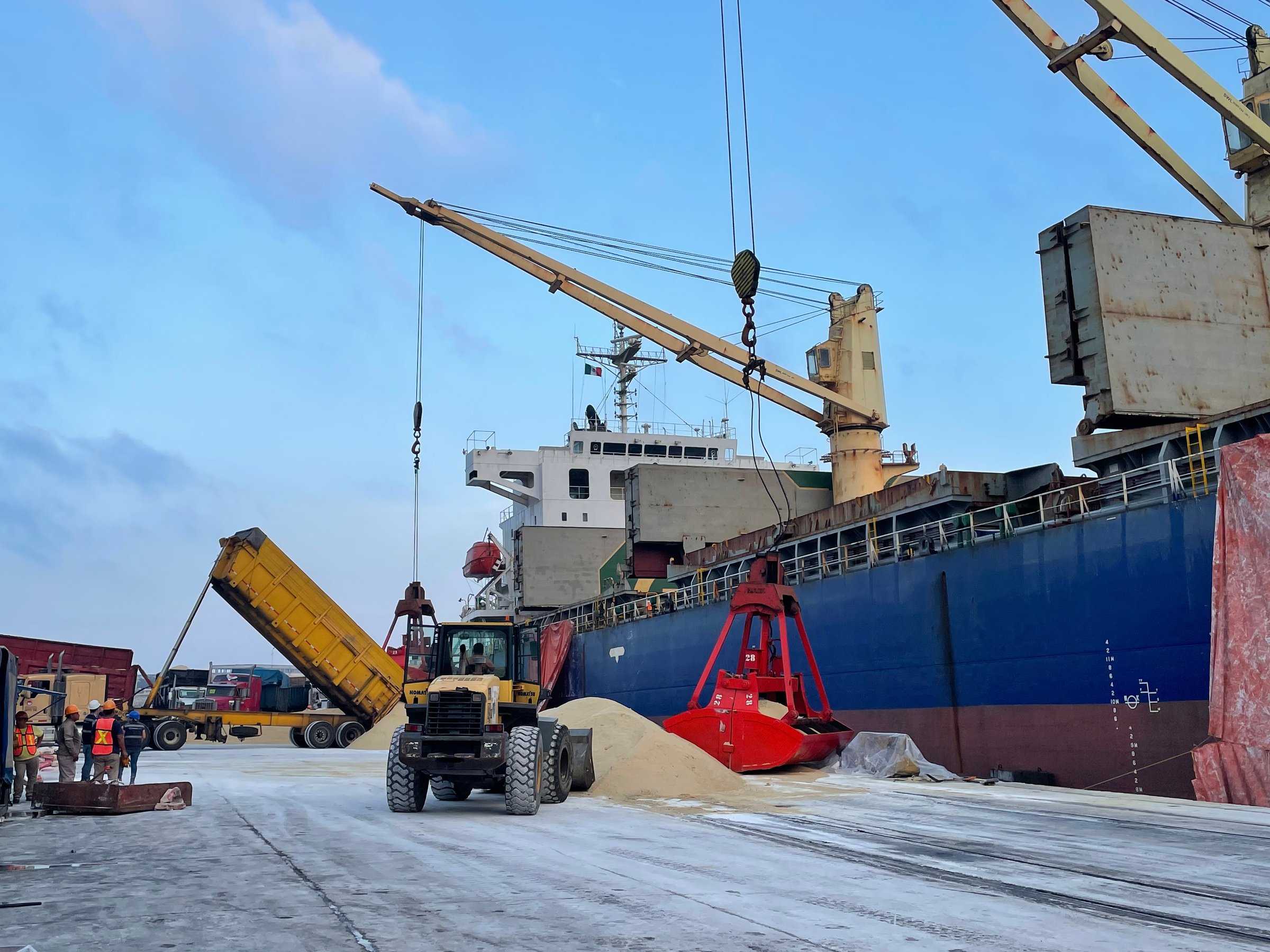 Cargo ship docked at port with crane unloading materials, alongside workers and a dump truck tilting its bed.
