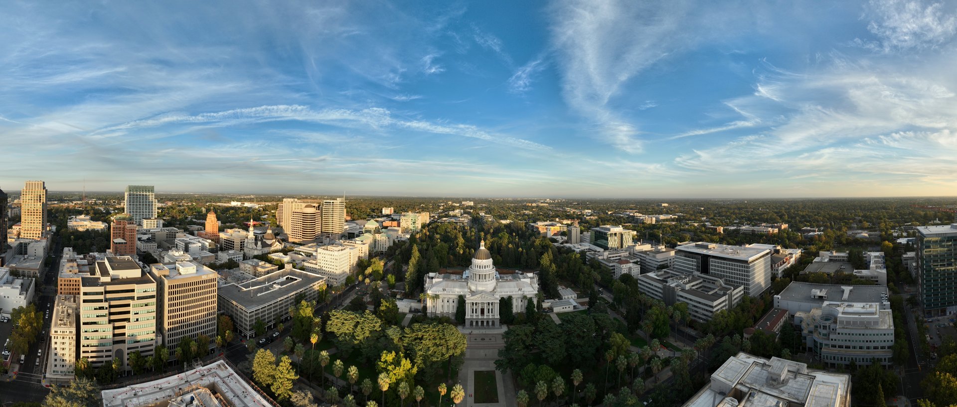 A panoramic aerial view of Sacramento's skyline, featuring the California State Capitol building, under a bright, partly cloudy sky.