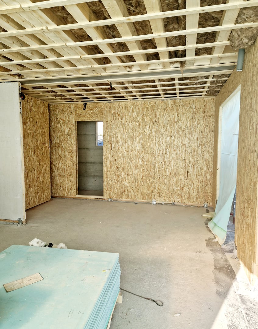 A partially constructed interior of a prefabricated wooden house, featuring OSB panel walls and an unfinished ceiling structure with visible insulation. The open doorway leads to another section of the house, showcasing the progress of modular home construction. The space is illuminated by natural light, emphasizing the texture of the engineered wood panels and the simplicity of the design.