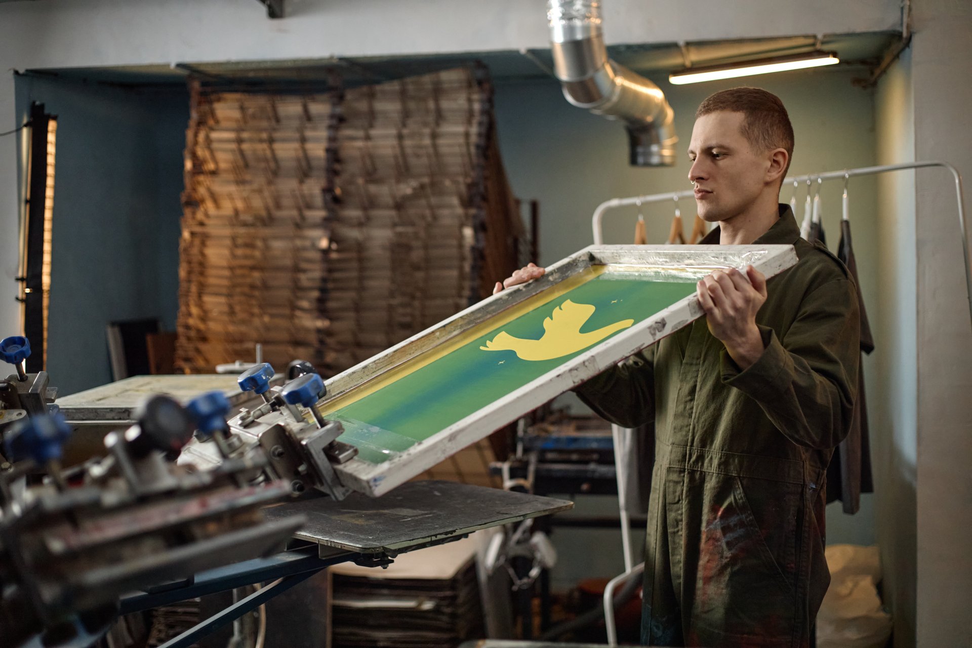 Caucasian young adult man screen printing yellow bird design onto fabric in workshop, holding large frame and focusing on precise alignment, industrial equipment visible in background