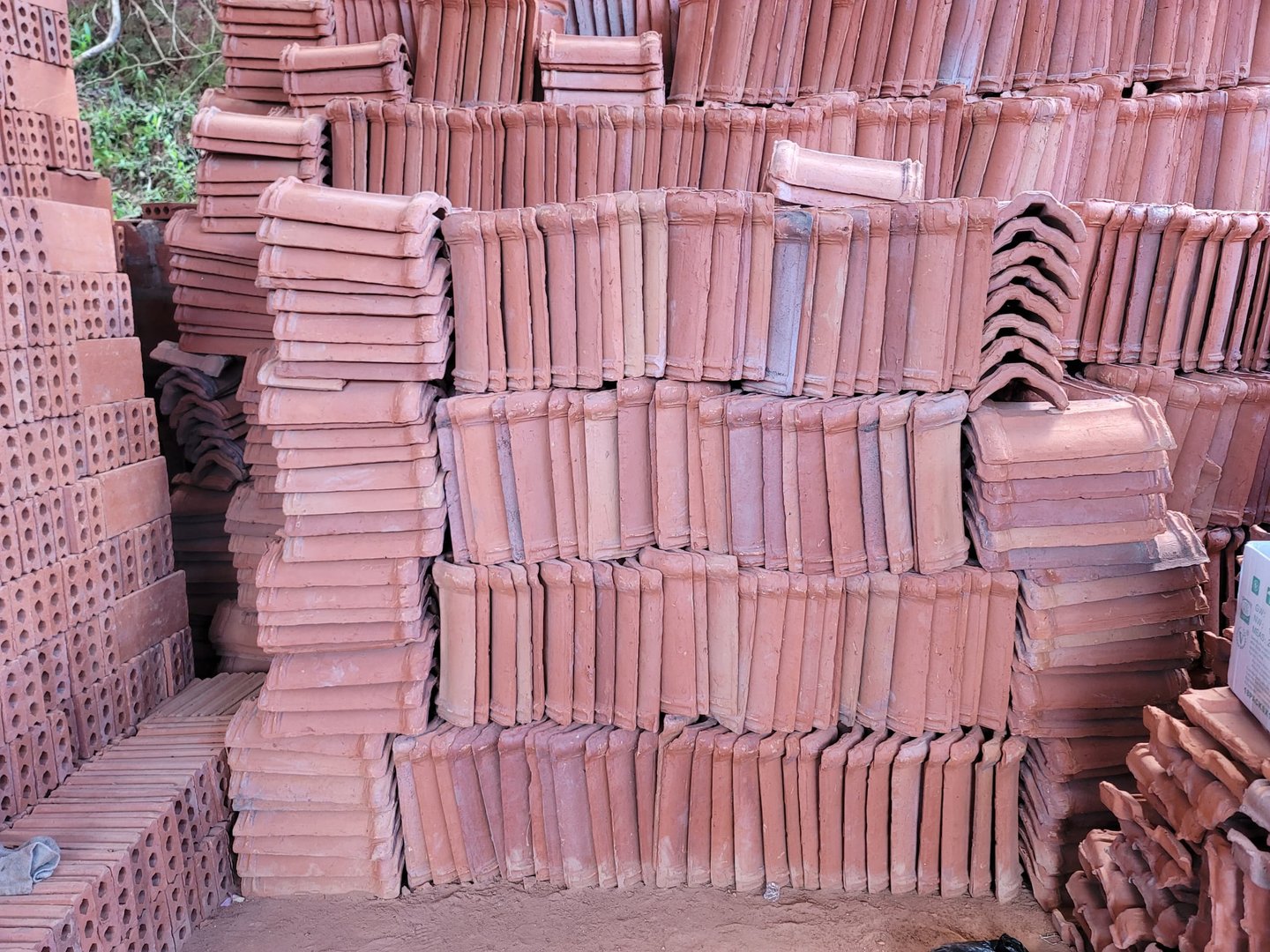 Stacks of rectangular terracotta roof tiles and bricks neatly arranged against a wall on a dirt floor.