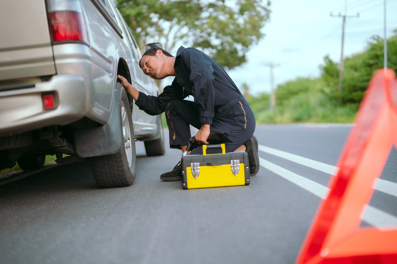 A mechanic kneels beside a vehicle on a rural road, inspecting its tire. A warning triangle sits on the side, indicating a breakdown as daylight fades in the background.