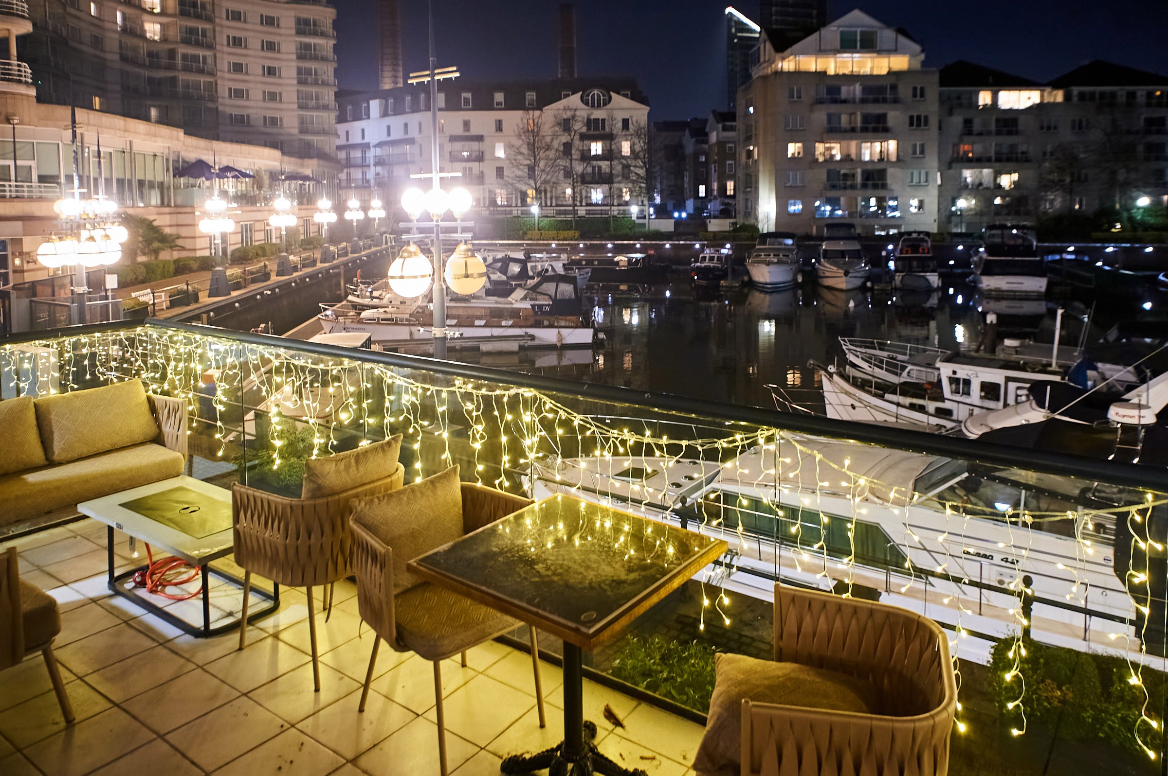 Night view of a marina with boats, festive string lights, and outdoor seating on a terrace overlooking the water.